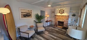 A living room with a fireplace, two armchairs, a surfboard, and a framed picture on the wall. - Most Photographed Bayfront-Amazing Sunset & Palm Trees on Exclusive Snug Harbor! (Stone Harbor)