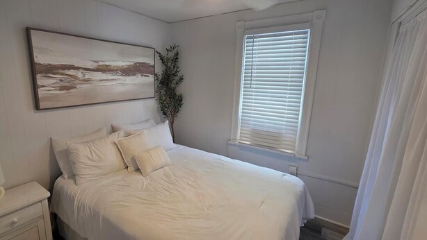 Ein Schlafzimmer mit einem Bett, einem Nachttisch, einer Lampe, einem Fenster mit Jalousien und einem Bild an der Wand.