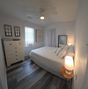 A bedroom with a bed, dresser, window, and a wall-mounted lamp. - Most Photographed Bayfront-Amazing Sunset & Palm Trees on Exclusive Snug Harbor! (Stone Harbor)