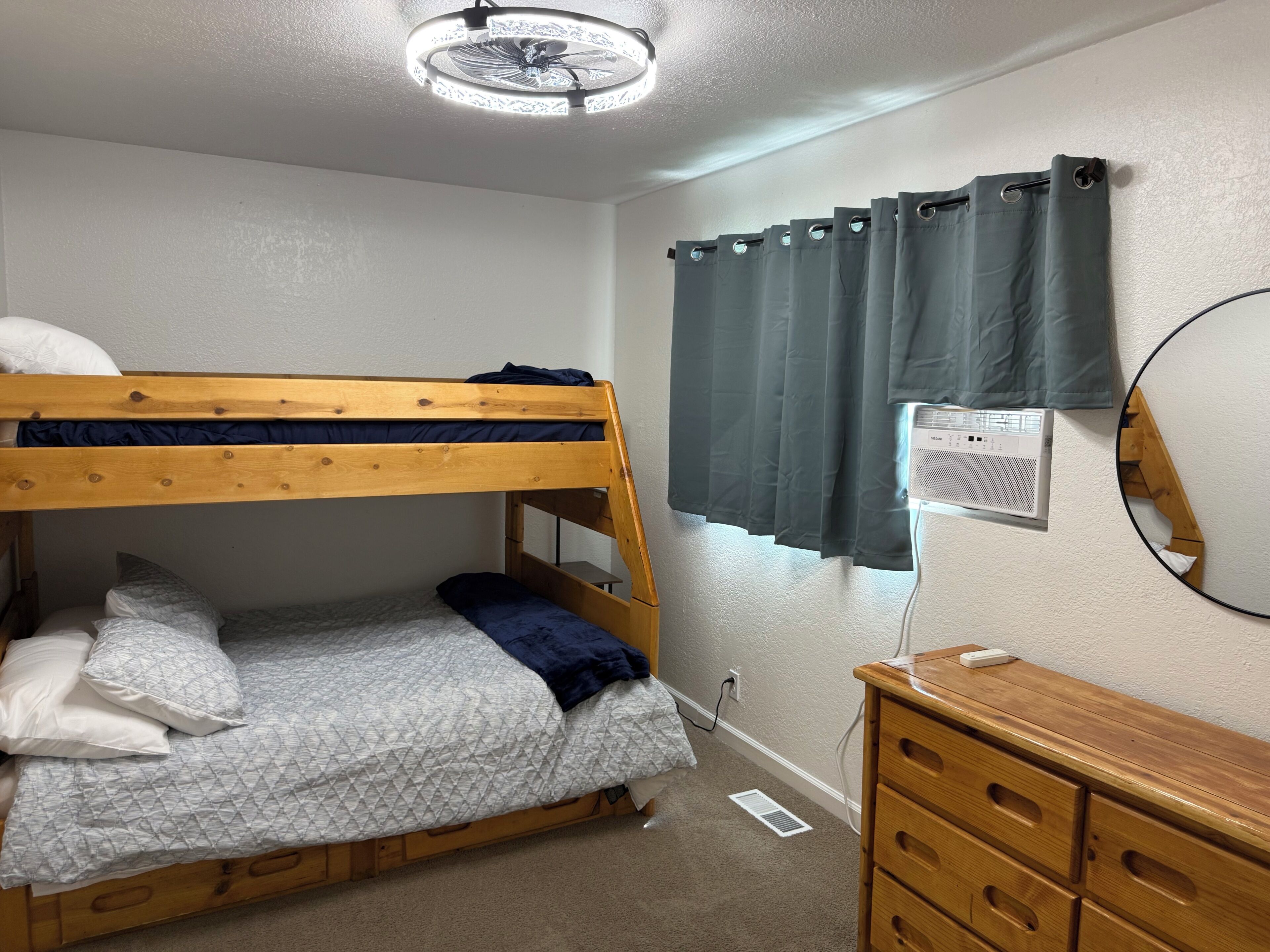 Un lit superposé avec une tête de lit, une commode avec des tiroirs, un radiateur fixé au mur et une fenêtre avec des rideaux.