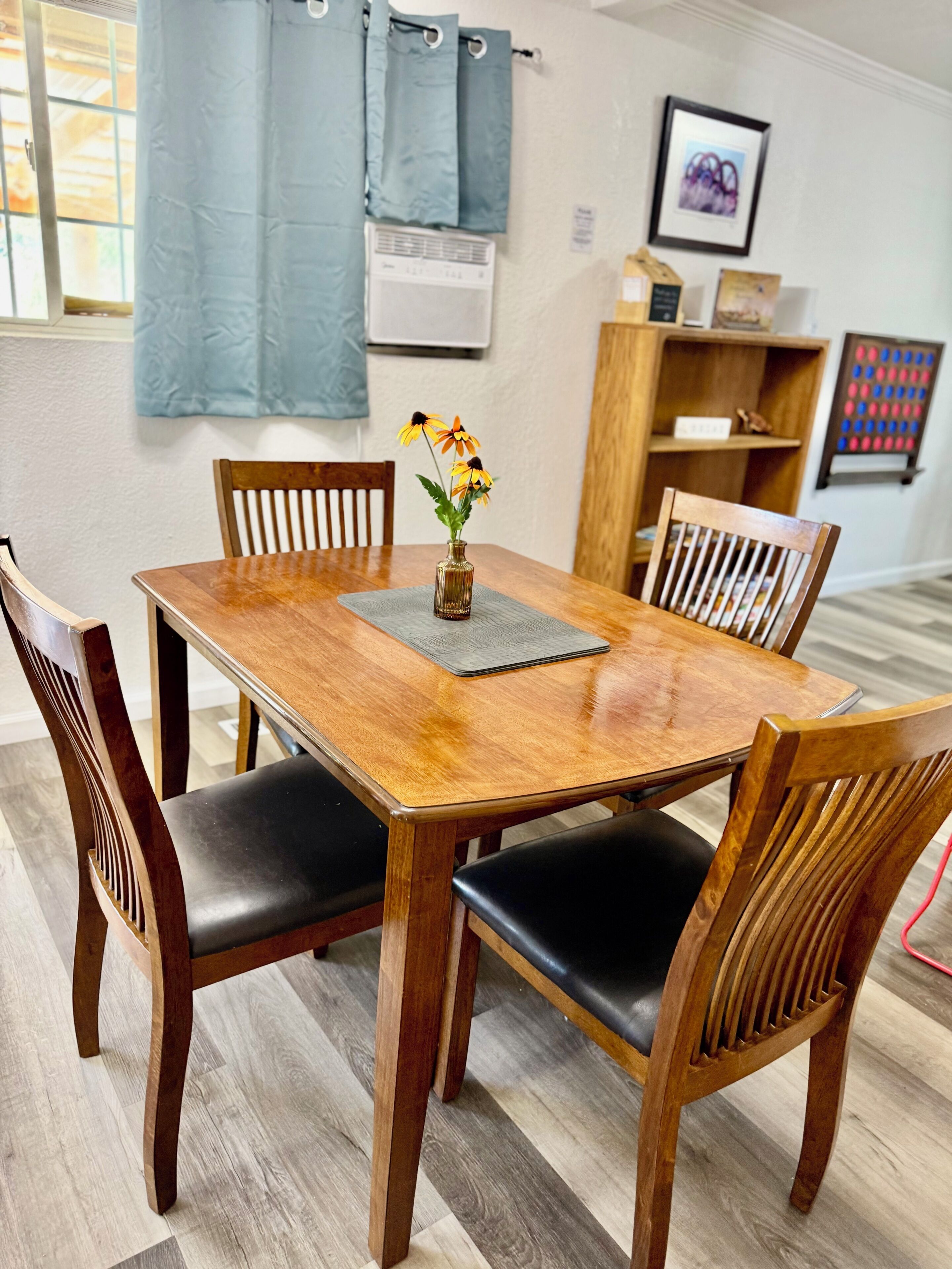 A wooden dining table with four chairs, a vase with flowers, and a small shelf with books and a picture frame.