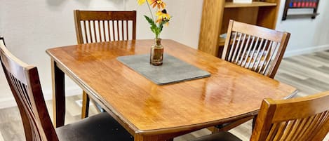 A wooden dining table with four chairs, a vase with flowers, and a small shelf with books and a picture frame.