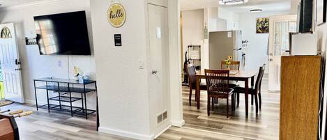 A spacious living area with a dining table, a flat-screen TV mounted on the wall, and a kitchen in the background.