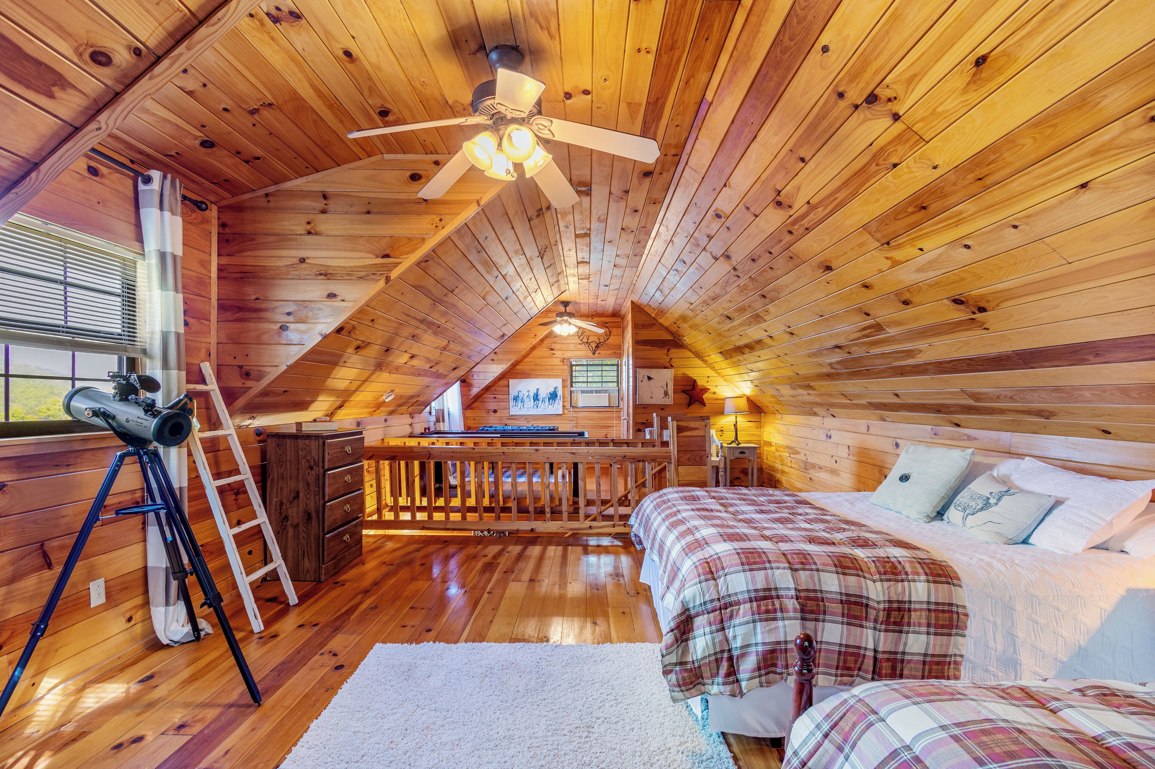 A cozy attic bedroom with wooden walls, a ceiling fan, a telescope, and a bed with plaid bedding.