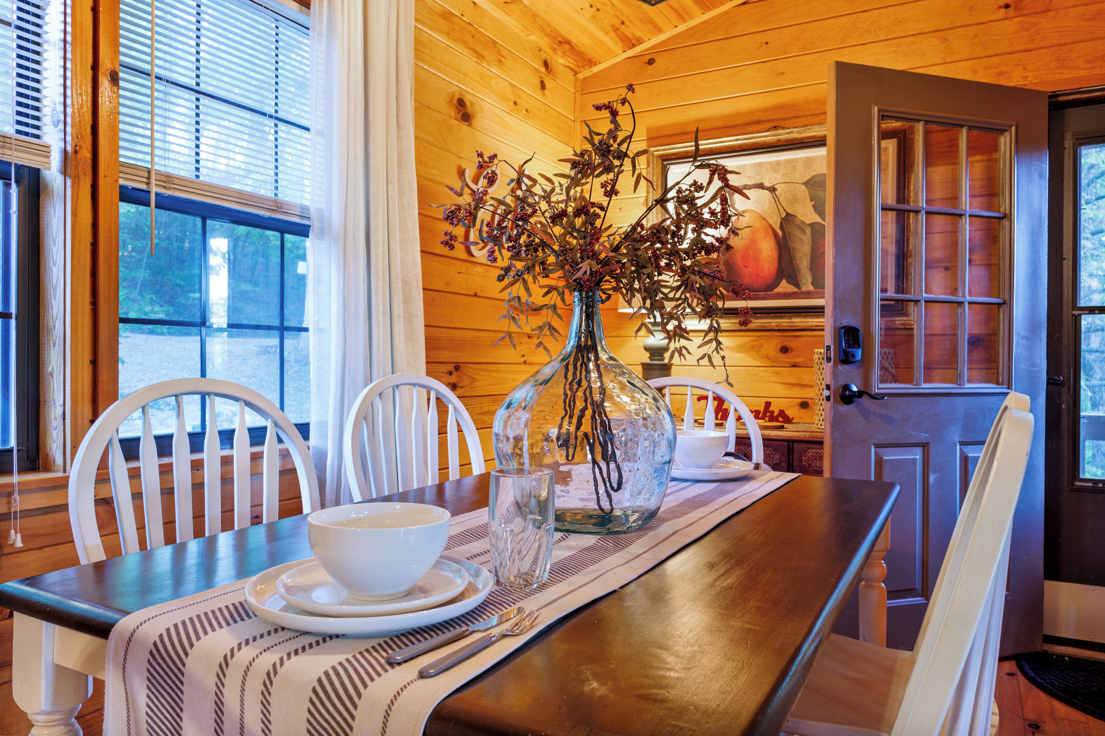 A wooden interior with a dining table set for two, a vase with dried flowers, and a door with a glass panel.