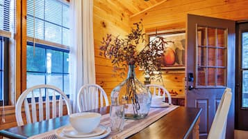 A wooden interior with a dining table set for two, a vase with dried flowers, and a door with a glass panel.