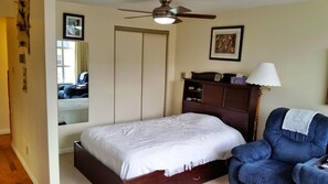 Un dormitorio con cama, una silla, armario, ventilador de techo y mesita de noche con lámpara.