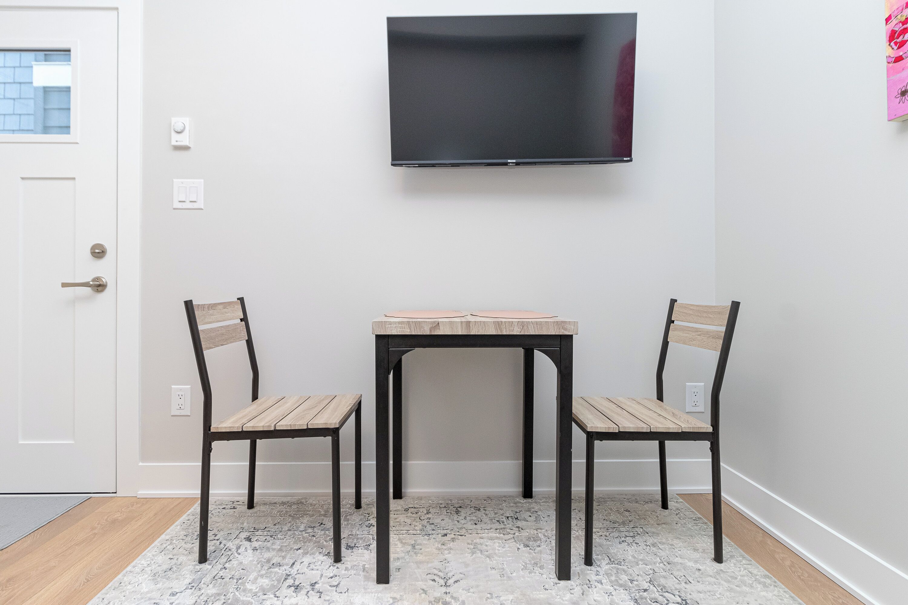 A room with a flat-screen TV mounted on the wall, a small wooden table, two chairs, and a patterned rug on the floor.
