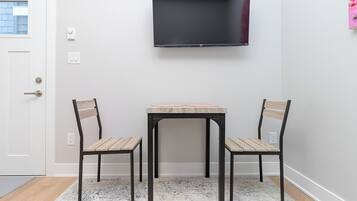 A room with a flat-screen TV mounted on the wall, a small wooden table, two chairs, and a patterned rug on the floor.
