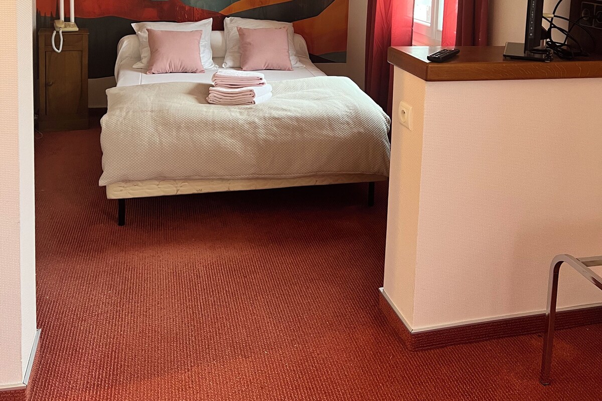Een hotelkamer met een bed, een bureau met een computer, een muur met een berglandschap en een raam met rode gordijnen.