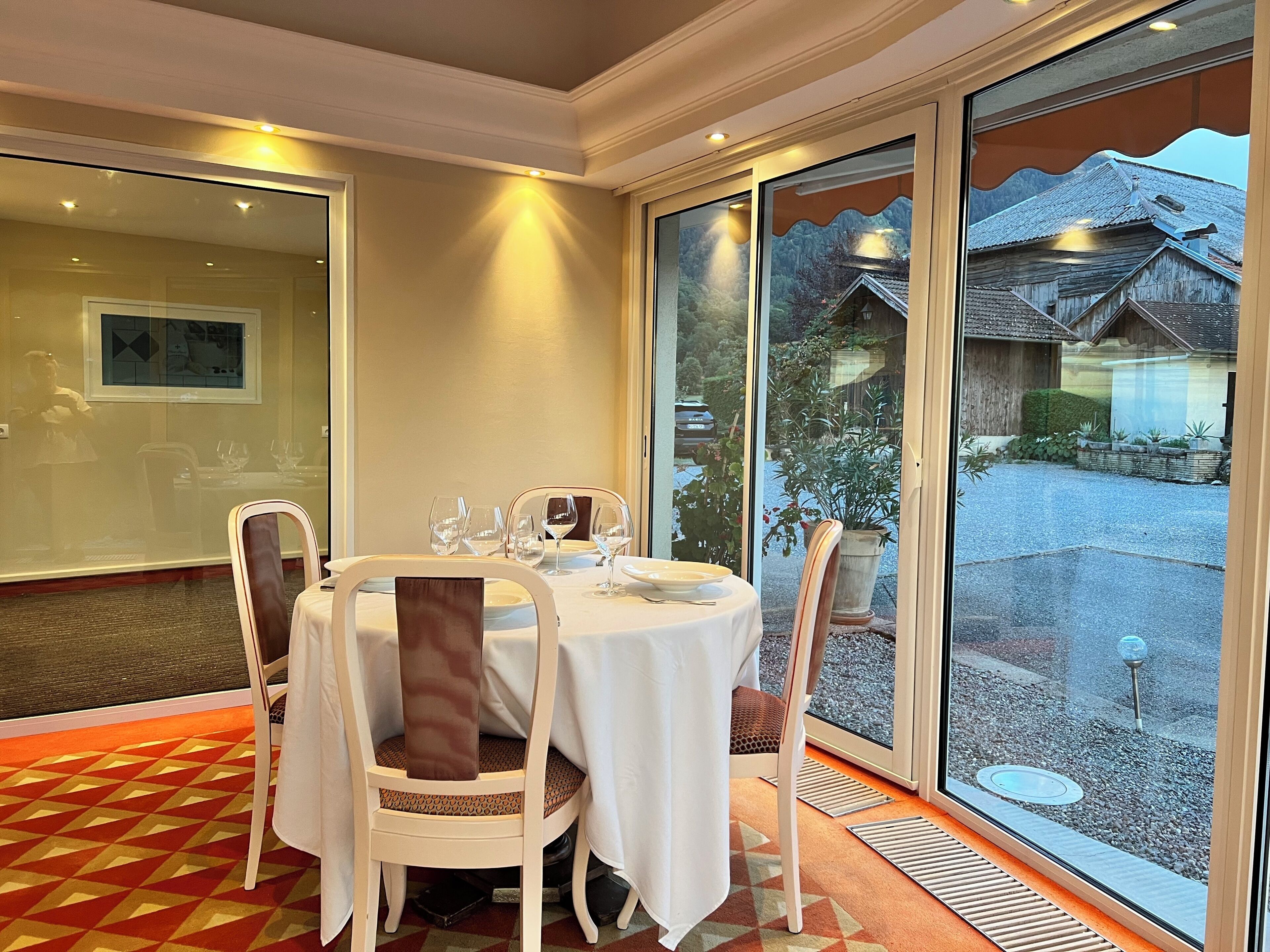 A dining area with a round table, chairs, and a view of a traditional building through large windows.