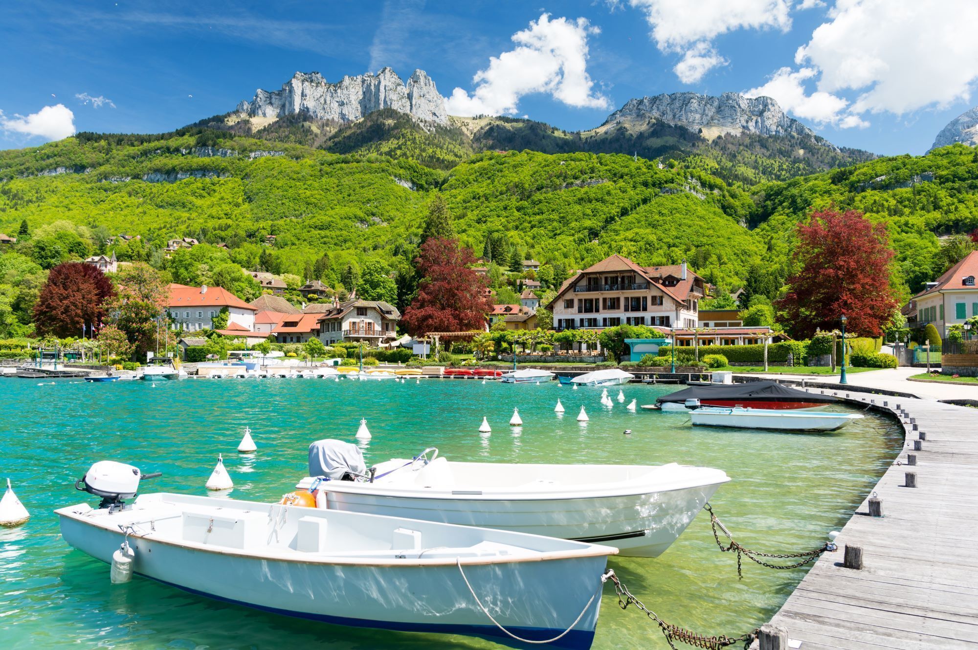 A marina with boats docked, a wooden pier, and buildings with red roofs in the background.