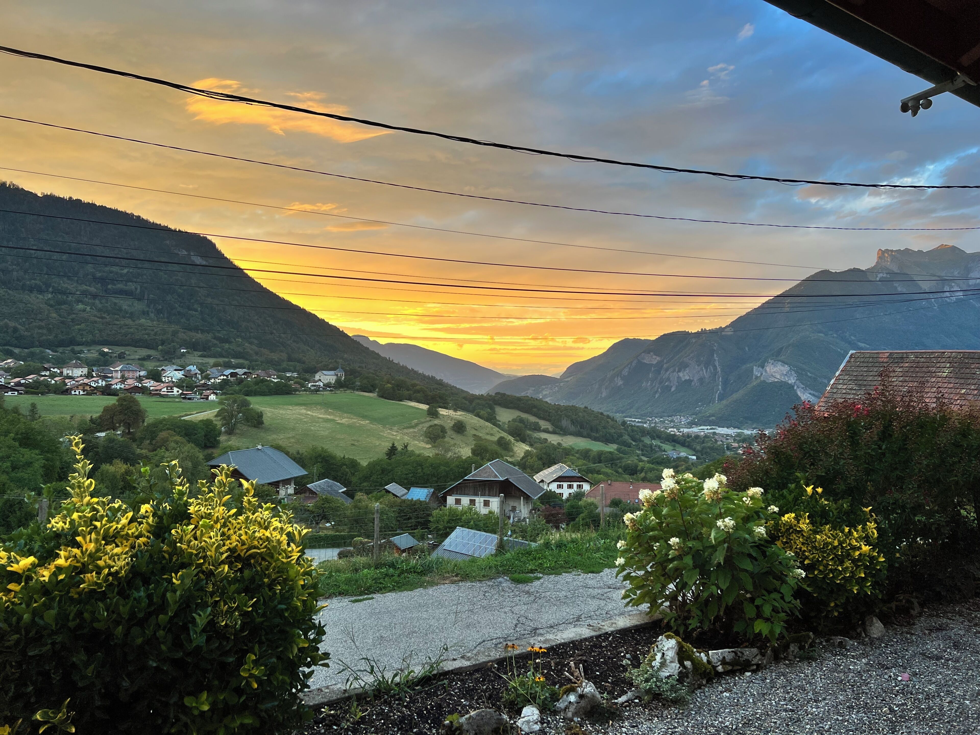 Paisagem rural com casas, um vale e montanhas sob um céu ao pôr do sol.