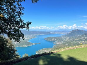 A panoramic view of a coastal landscape with a clear blue lake, surrounding hills, and a distant mountain range. - VICC (Faverges-Seythenex)