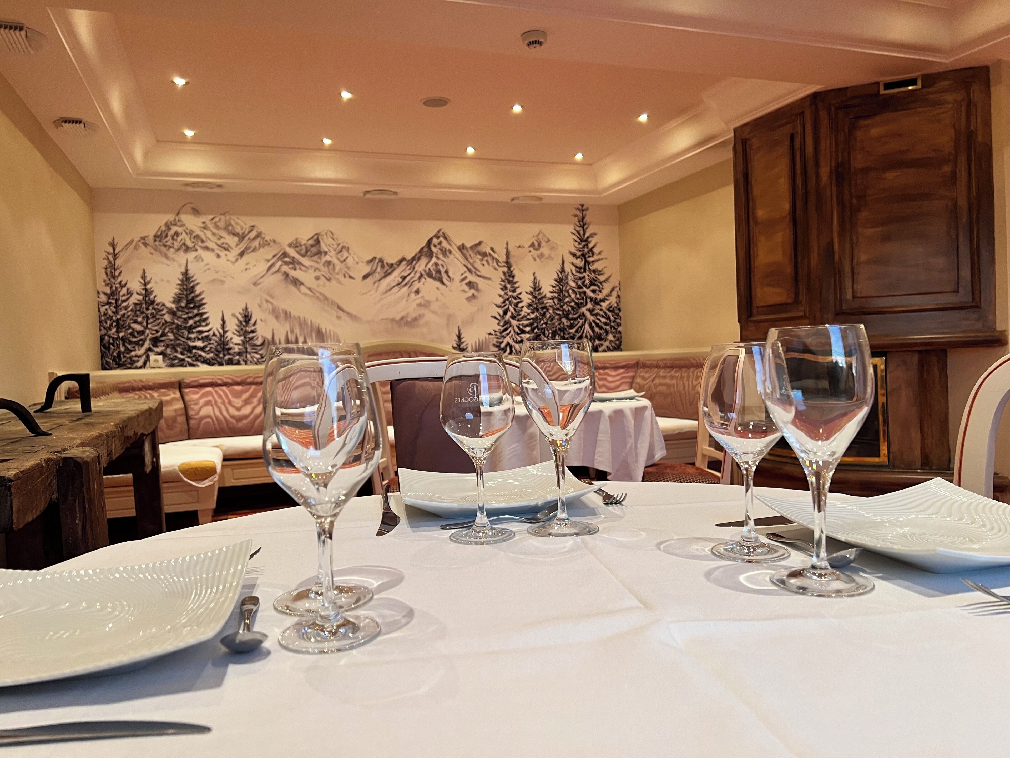 Uma sala de jantar com uma mesa posta para quatro pessoas, com taças de vinho e pratos, e um mural com tema de montanhas na parede.