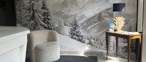 A room with a mountain mural, a white chair, a small table with a lamp, and a tiled floor.