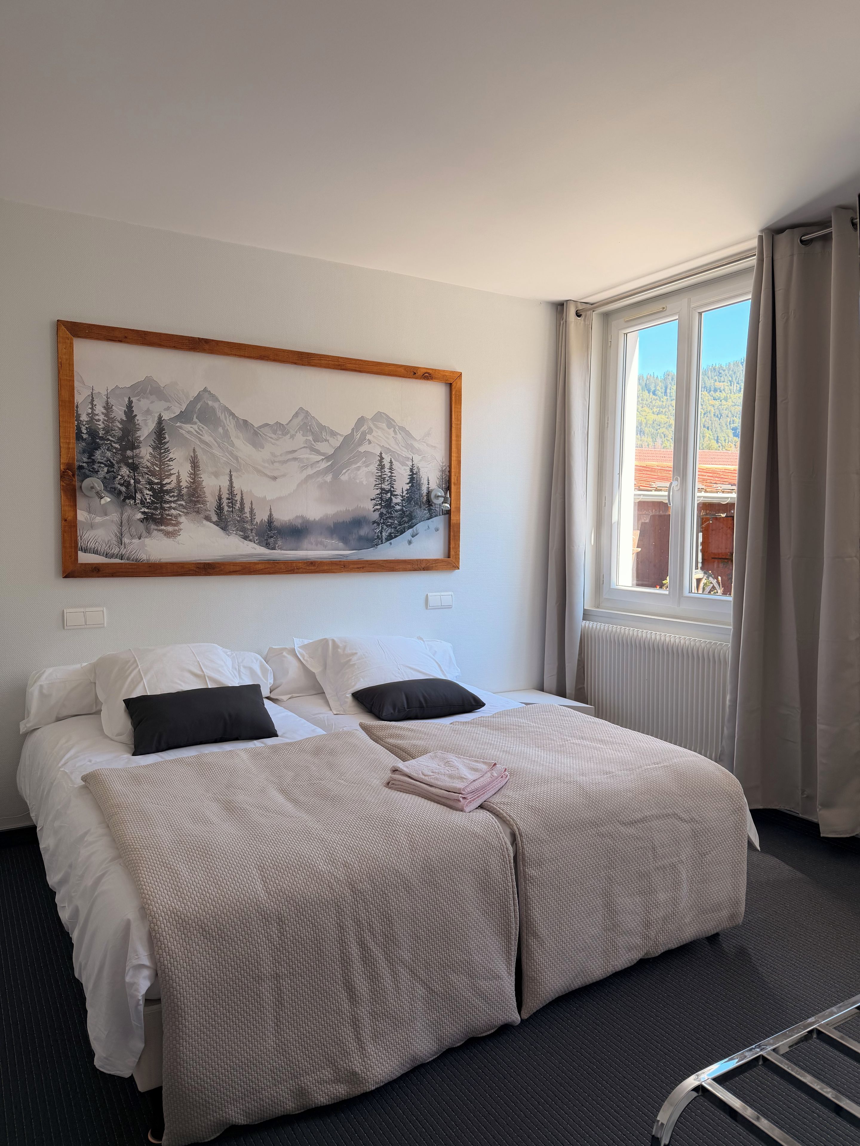 A hotel room with a bed, a painting of a mountain landscape, a window with curtains, and a radiator.
