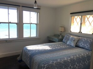 Un dormitorio con una cama, dos ventanas, una mesita de noche y una lámpara.