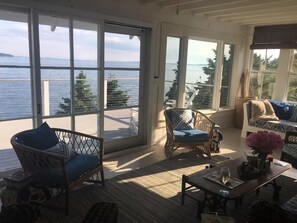 Una habitación iluminada por el sol con ventanas amplias que dan a un cuerpo de agua, amueblada con sillas de mimbre y una mesa de madera.