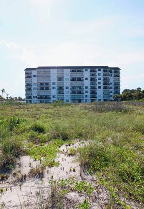 A multi-story building with a flat roof, surrounded by a natural landscape with sparse vegetation. - Step into the Beach Winds / Ocean Breeze Patio (Cocoa Beach)