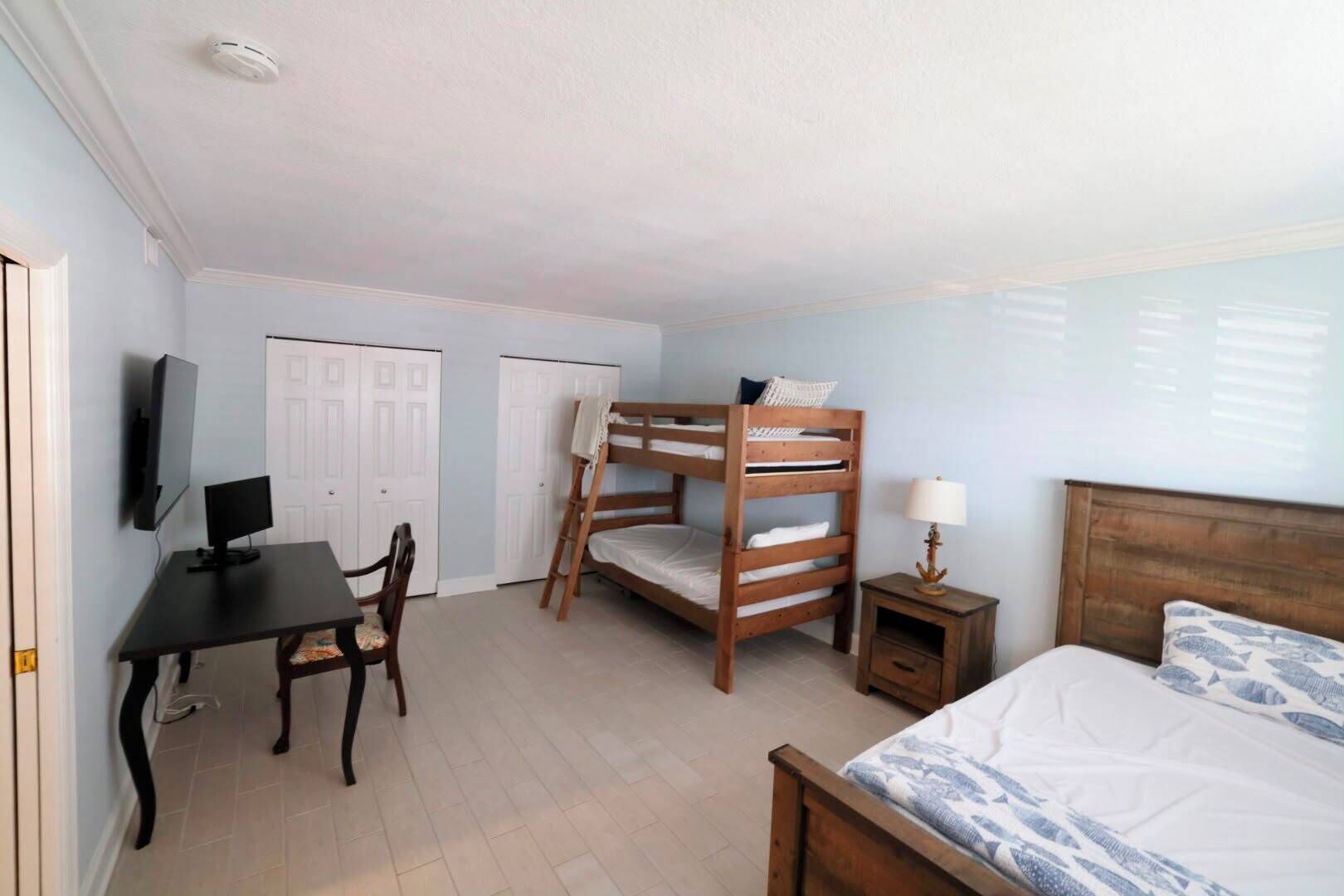 A room with bunk beds, a desk with a computer, a chair, a bedside table with a lamp, and a bed with a patterned blanket.