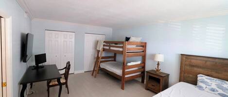 A room with bunk beds, a desk with a computer, a chair, a bedside table with a lamp, and a bed with a patterned blanket.