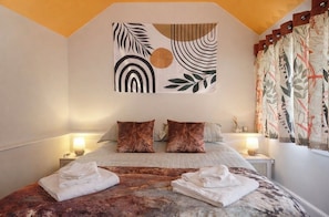 A hotel room with a bed, two pillows, a wall tapestry, a nightstand with a lamp, and a window with curtains. - Garden Galley Nook | Coast & Countryside Haven (Sproatley)