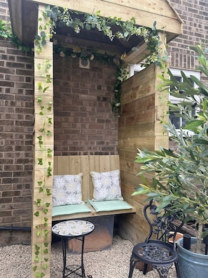 A wooden bench with cushions, a small table, and potted plants in a brick-walled outdoor seating area. - Garden Galley Nook | Coast & Countryside Haven (Sproatley)