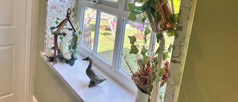 Ein Fenster mit dekorativer Gardine, eine kleine Ablagefläche mit einer Entenfigur und eine hängende Makramee-Pflanze.