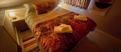 Una cama bien tendida con una colcha estampada en rojo y naranja, dos almohadas y dos toallas dobladas sobre la cama.