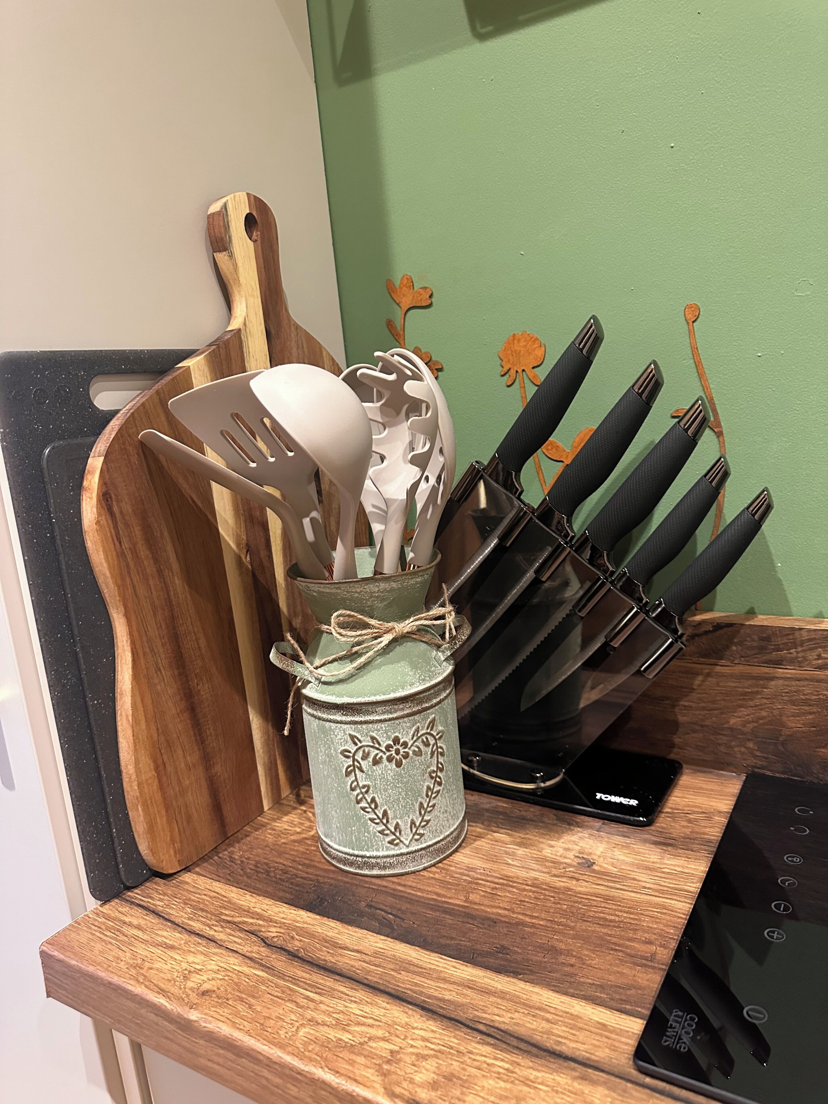 Una encimera de cocina con una tabla de cortar de madera, un recipiente metálico con utensilios y un dispositivo electrónico negro.