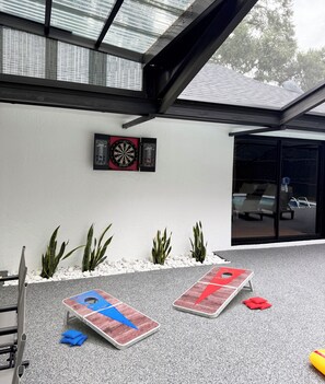 A covered outdoor area with a dartboard and two shuffleboard tables. - Modern Pool Retreat • Outdoor Kitchen • Fire Pit • Pets (Palm Coast)
