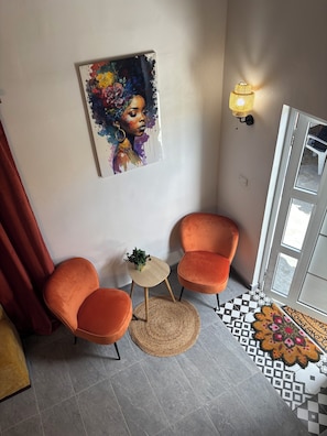 Ein Zimmer mit zwei orangefarbenen Sesseln, einem kleinen runden Tisch mit einer Pflanze, einem farbenfrohen Gemälde an der Wand, einer Wandleuchte und einem gemusterten Teppich.