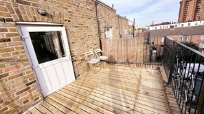 A wooden deck with a door, two chairs, and a small table. - Bright Chelsea Studio w/ Private Balcony | King Bed | Sleeps 2 (London)