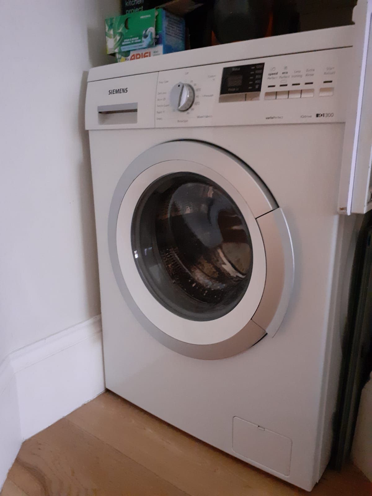 Een witte Siemens wasmachine met bedieningspaneel en glazen deur, geplaatst in een hoek met een houten vloer.