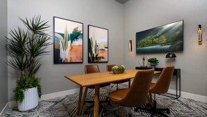 A modern dining area with a wooden table, four chairs, abstract paintings, a large TV, and a potted plant.