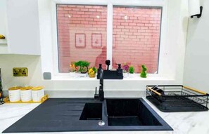 A kitchen with a black sink, a window with a brick wall view, and a shelf with plants and kitchenware. - Cozy and Beautiful 3-Bed House in Manchester!
Sleeps 6 Comfortably! (Salford)