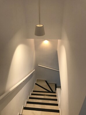 Una escalera con barandilla, una lámpara arriba y una pared blanca.