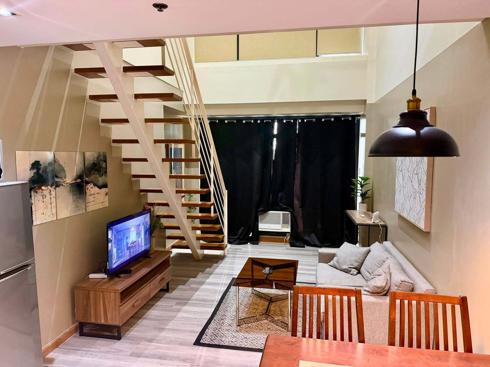 A modern living room with a wooden staircase, a flat-screen TV, a sofa, and a dining area.