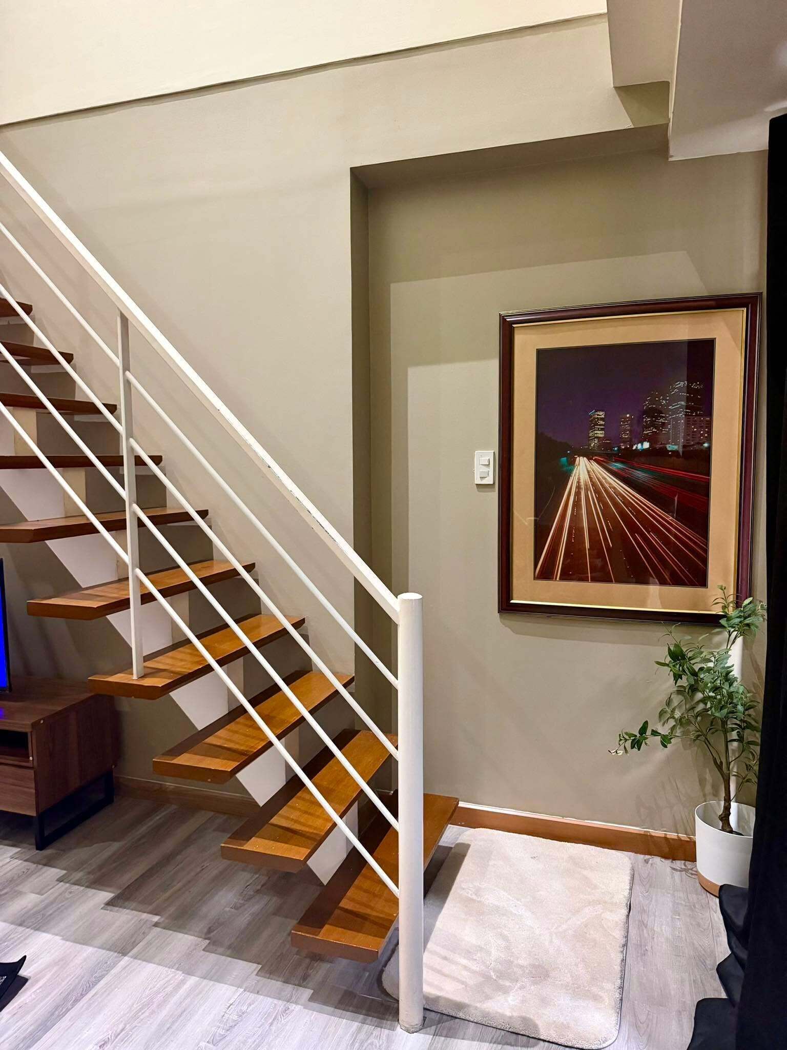 Una escalera con peldaños de madera y barandillas blancas, un cuadro enmarcado en la pared y una planta en maceta.