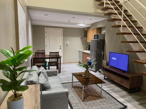Una sala moderna con sofá, mesa de centro y televisor. Se ve una escalera a la derecha y una cocina al fondo.