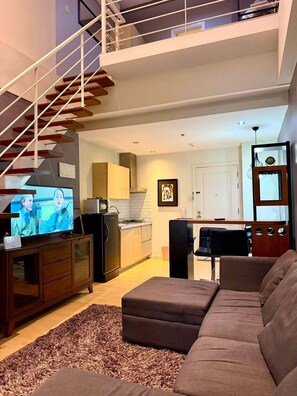 A modern living room with a staircase, a flat-screen TV, a sofa, and a kitchen in the background. - The Gramercy Residences  (Makati)