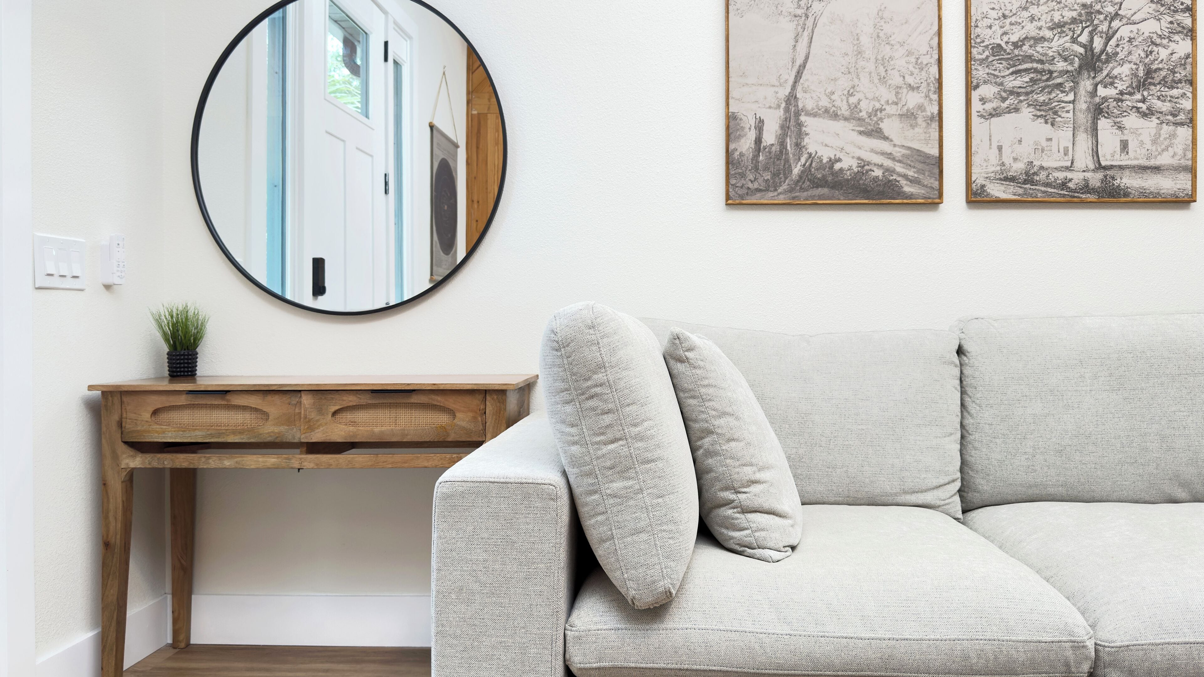 A living room with a round mirror, a wooden desk, a light-colored sofa, and two framed pictures on the wall.