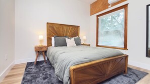 Un dormitorio con cabecera de madera, cama con ropa de cama gris, dos mesitas de noche con lámparas y una ventana con persianas.