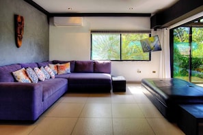 A modern living room with a sectional sofa, a flat-screen TV, and a large window with a view of greenery. - Casa Siempre Verde Ocean View (Nosara)