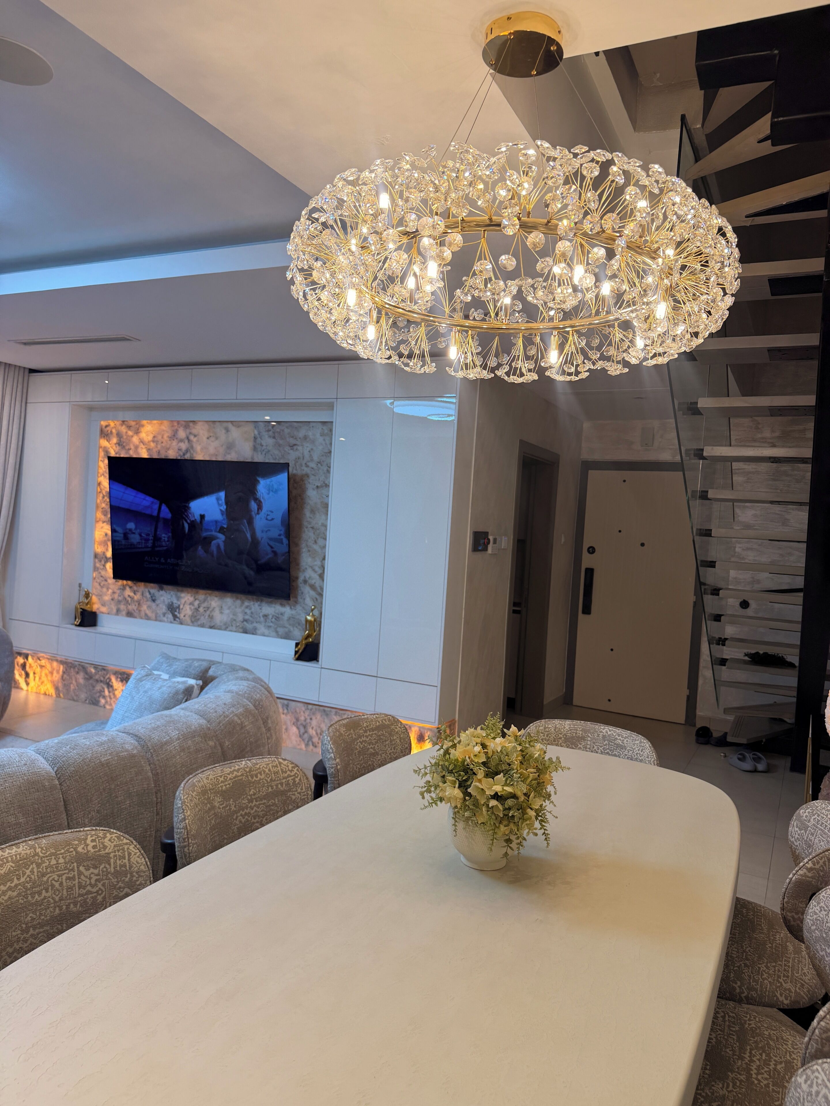 Una sala moderna con una lámpara de araña grande, un televisor de pantalla plana y una zona de comedor con un florero.