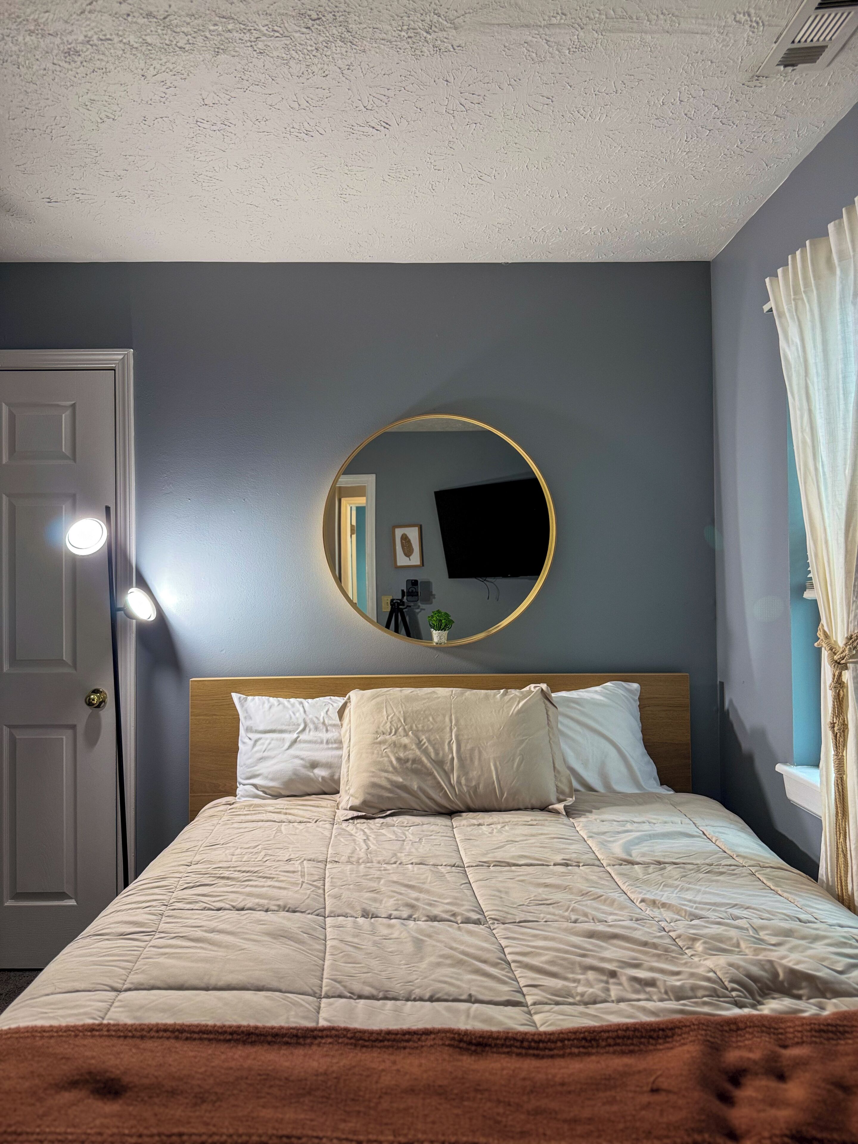 A bedroom with a bed, a round mirror, a door, and a window with curtains.