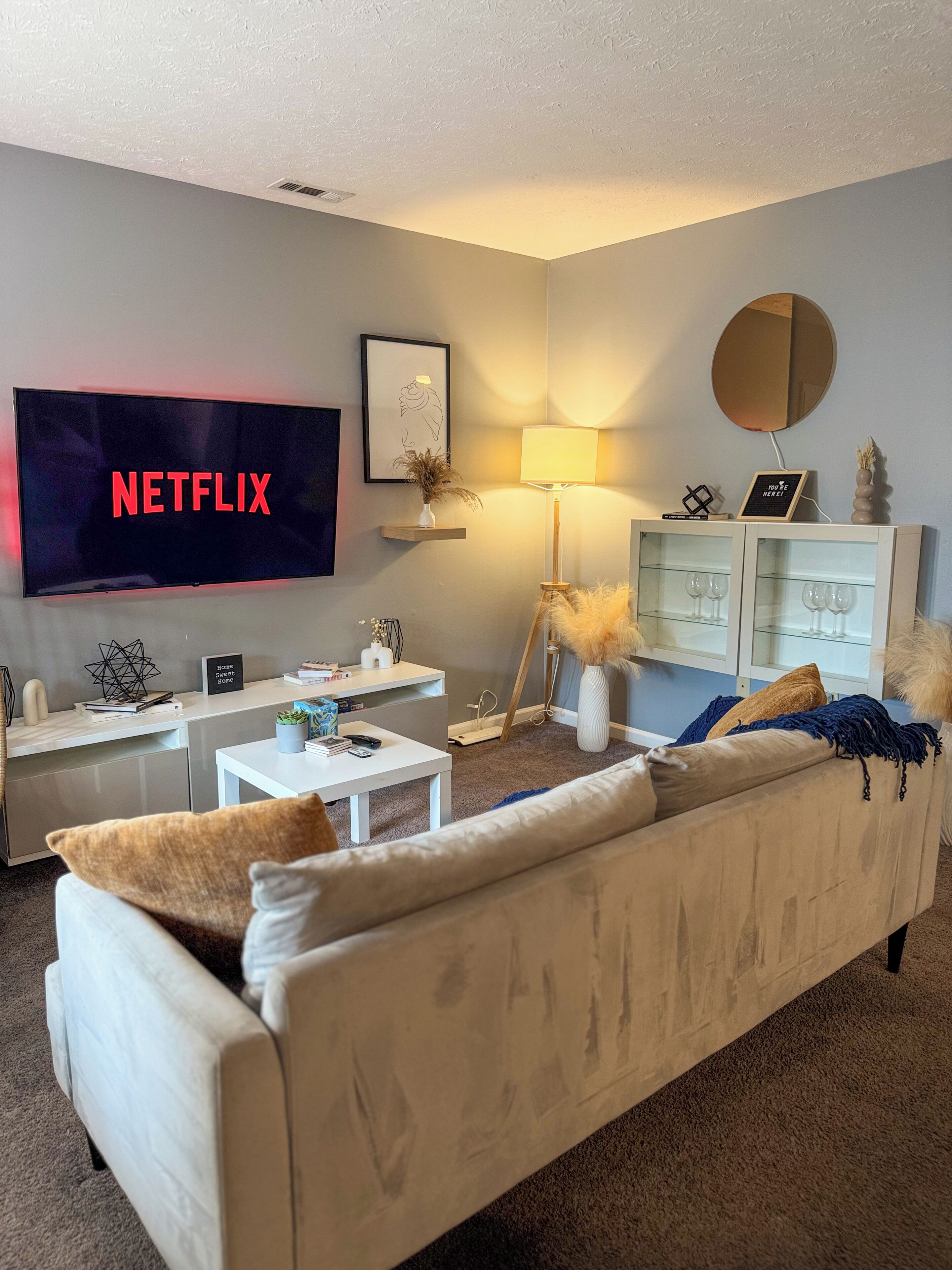 A living room with a flat-screen TV displaying 'NETFLIX,' a white sofa, a coffee table, a floor lamp, and a cabinet with glass doors.