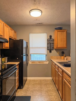 A kitchen with wooden cabinets, a black refrigerator, a stove, and a window with blinds. - Charming 2BR Indianapolis Retreat for Extended Stays (Indianapolis)