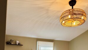 Ein Schlafzimmer mit einem Bett, Kissen, einem Fenster mit Jalousien, einer Pendelleuchte und einem kleinen Regal mit Dekoration.
